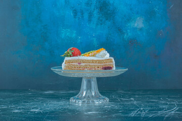 Fruit topped cake slice on a glass pedestal on blue background