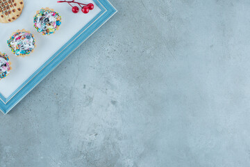 Candy cupcakes, a cookie and a christmas berry decoration on a board on marble background
