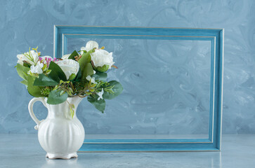 Empty picture frame behind a bundle of white roses in a vase on marble background
