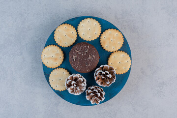 Biscuits around a chocolate cake on a blue board with pine cones on marble background