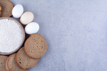 Sliced black bread and eggs around a bowl of flour on marble background