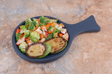 Serving pan with fried zucchini slices and a mixed vegetable salad on marble background