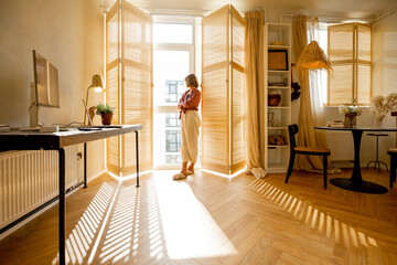 Young woman stands alone by the window blinds at cozy and sunny living room of modern apartment in...