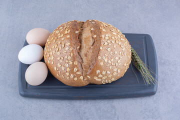 Three eggs, a loaf of bread and a single wheat stalk on a board on marble background
