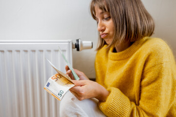 Warmly dressed woman counting money while sitting near radiator at home. Concept of expensive...