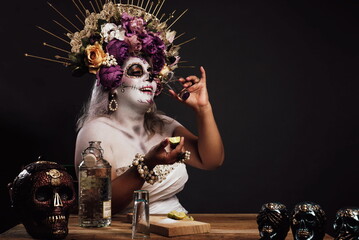 Portrait of woman in catrina makeup wearing wedding dress drinking tequila in a bar. Colorful skulls on the table. - obrazy, fototapety, plakaty