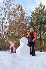 Portrait of little girl sitting on snow and mother making snowman on backyard in evening with rowan and fir trees in background. Parents spending time with children.
