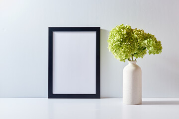 Mockup with a black frame and green flowers in a vase on a light background. Empty poster frame mockup for presentation design, text, lettering