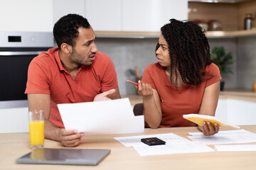 Upset millennial black wife and husband, with tablet and documents pay bills and taxes, quarrel in kitchen interior