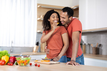Obraz na płótnie Canvas Cheerful millennial black husband hugging lady, woman cutting organic vegetables at salad and feeding man