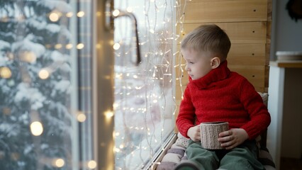 Naklejka premium A little boy in a red sweater sits on the windowsill and watches as it snows outside the window. The boy is waiting for the New Year holidays.
