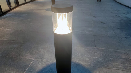 Low street lamp. On the concrete pavement stands a low metal lantern with a lamp at the top. The...