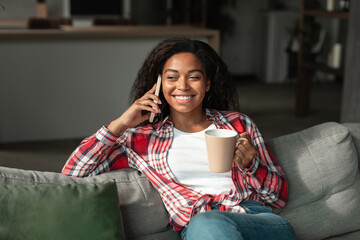 Fototapeta Smiling millennial black female with cup of drink, enjoy free time, talk on phone, sit on sofa in living room obraz