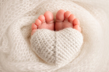 The tiny foot of a newborn baby. Soft feet of a new born in a white wool blanket. Close up of toes, heels and feet of a newborn. Knitted white heart in the legs of a baby. Macro photography. 