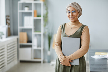 Minimal waist up portrait of young Middle Eastern businesswoman smiling at camera in office setting and holding laptop, copy space