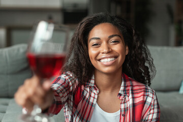 Glad millennial african american lady with glass of wine cheers at camera, have video call in living room interior