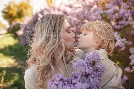 A Little Stylish Boy  With Mother  Walks Among Flower Trees In Spring And Mom  Holds A Kid And Hugging Outdoor In The Nature