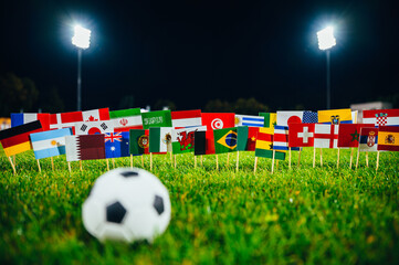 Football ball on green grass. 32 flags of all nations. Dark background, Stadium in night. Artificial light. Black edit space