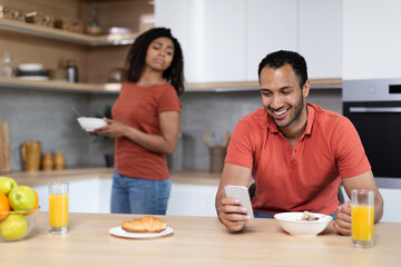 Jealous millennial black wife looks at phone husband, chatting by phone at breakfast in kitchen interior