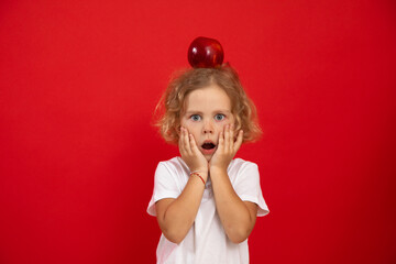 Portrait of shocked little girl with short curly fair hair, red apple on head wearing white T-shirt, holding cheeks.