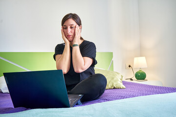 Caucasian woman with hands on face looking at computer from bedroom