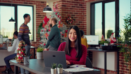 Portrait of employee sitting at desk to work on laptop, sending email before celebrating winter holiday in office. Working on computer in space with christmas tree and seasonal decor.