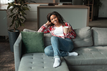 Cheerful millennial african american lady with cup of favorite drink, enjoy free time and comfort, sit on sofa