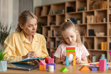 Female psychotherapist working with little girl at office, sitting by preschooler playing with bricks, taking notes - obrazy, fototapety, plakaty