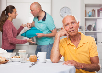 Elderly man sitting at table, unhappy family couple quarrelling on backgroud