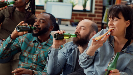 Diverse friends making toast with beer bottles and watching movie on tv channel. Having fun with cinema film on television program, saying cheers at social gathering. Handheld shot.