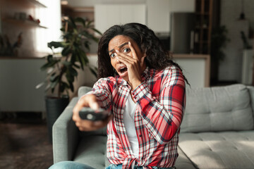 Afraid millennial african american woman with remote control sits on sofa and covers face with hands