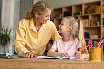 Mom and her little daughter sitting at desk and smiling to each other, mother helping her child with school homework