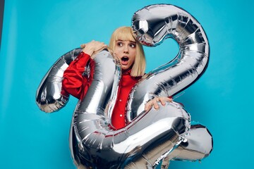 a surprised, woman in a red shirt stands on a blue background and holds inflatable balloons in the shape of the number twenty-two in silver color, hugging them with her hands, her mouth wide open