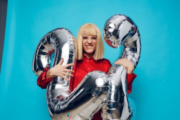 a happy, enthusiastic woman in a red shirt stands on a blue background and holds inflatable balloons in the shape of the number twenty-two in silver color, smiling broadly and hugging the balloons