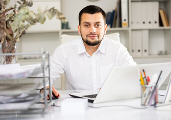 Cheerful man office worker sitting at desk and using laptop during his workday.