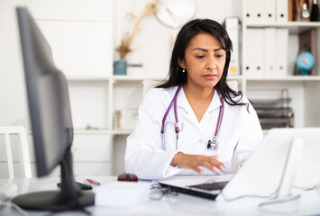 Latin american woman experienced physician filling up medical forms on laptop while sitting at...