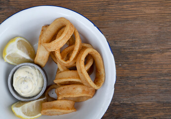 Delicious fried seafood. Closeup view of fried squid rings with lemon and a dipping sauce in a white bowl on the wooden table.