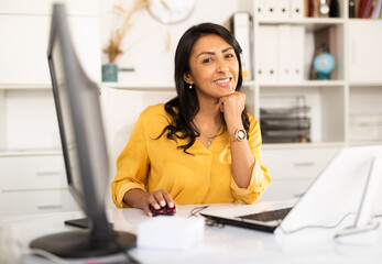 Portrait of confident smiling latin american female office employee during daily work with computer