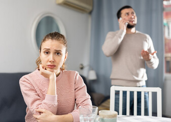 Frustrated offended young woman sitting at table at living room while her boyfriend talking on...
