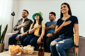 Multiethnic group of friends listening and singing Brazilian national anthem before watching sports championship on TV together at home - obrazy, fototapety, plakaty