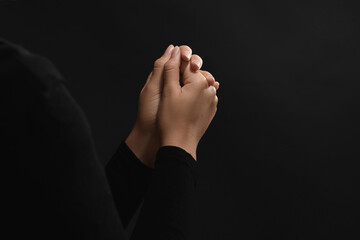 Woman holding hands clasped while praying against black background, closeup. Space for text