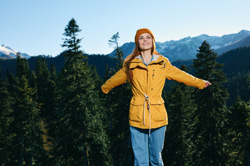 Woman with red hair hiker standing on the mountain hands up happiness overlooking snowy mountains and trees in yellow raincoat and cap travel autumn and hiking in the mountains in the sunset freedom