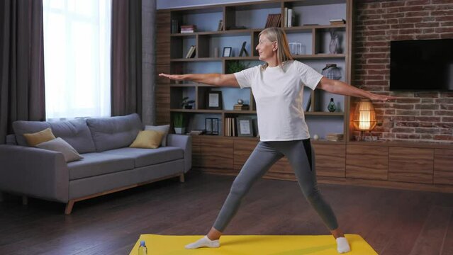Gray-haired happy mature woman doing gymnastics and yoga at home. Active and healthy lifestyle for pensioners. Fitness, stretching, relaxation, well-being of the elderly.