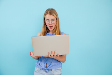 Portrait of surprised teenage girl wearing blue shirt, glasses, holding looking at screen of laptop on blue background.
