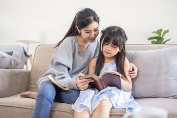 Obraz premium Happy asian mom holding her daughter playing and reading E-book on sofa at home.little girl hugging her mother smile and love having fun at home.Mother day concept