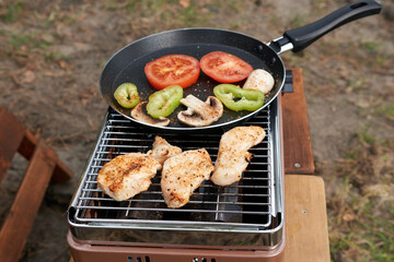 Portable gas grill that cooks chicken fillets and vegetables in a pan. The concept is a picnic in nature.