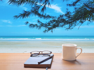 On the brown table, there are white coffee cup, pencil, diary (black notebook). 
The front overlooks sea, blue sky white clouds and top with green tree branches and leaves. - Powered by Adobe