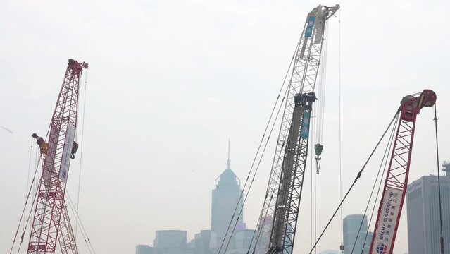 Numerous cranes stand and work on a construction site as part of an engineering redevelopment commercial project while the Hong Kong financial district skyline is in the background.