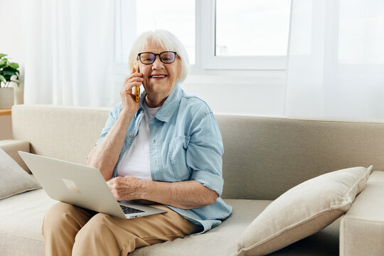 a happy, pleasant elderly lady with white hair is sitting on a cozy sofa in a bright apartment holding a laptop on her lap, holding it with her hand, laughing loudly with happiness