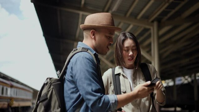 Couple Young Asian Traveler Tourist By Train Choose A Trip And Booking Train Queue By Smartphone. Two Backpacker At Platform Train Station. Happy Traveller At Railway On Vacation Holiday Weekend.